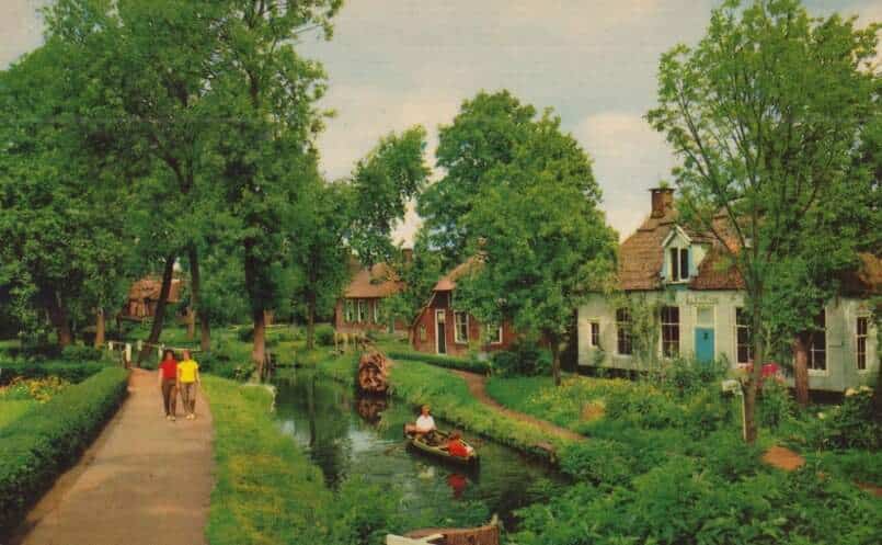 Archieffoto van een zomers dorpsbeeld in Giethoorn met gracht, wandelpad, erfbeplanting en een bootje dat rustig voorbijvaart.