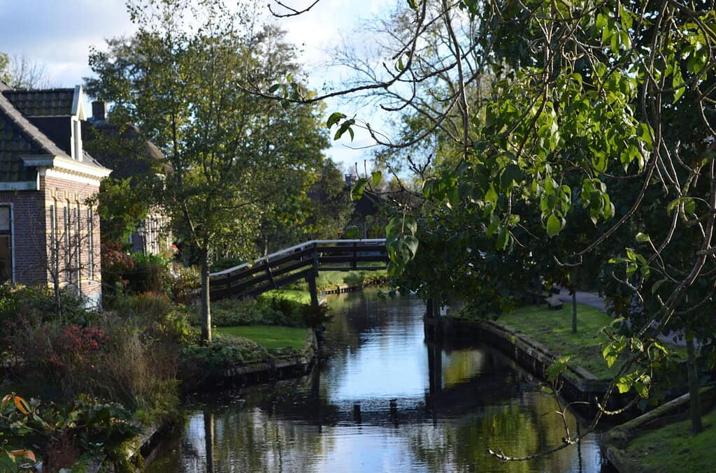 Houten brug in Giethoorn tussen bomen, struiken en woningen langs een smalle gracht.